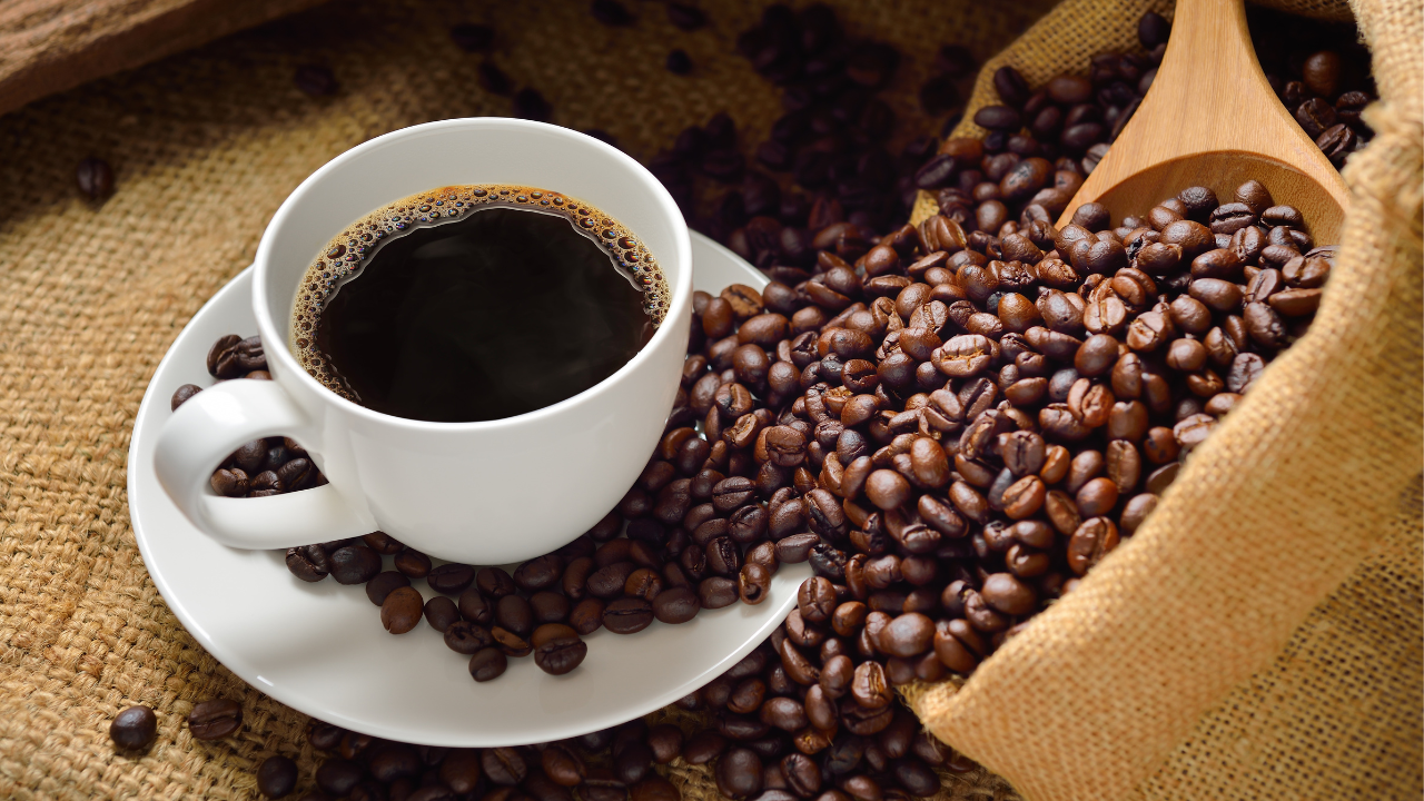 Picture of white cup and saucer filled with black coffee surrounded by coffee beans pouring out from a burlap bag
