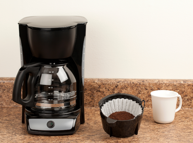 Picture of a coffee filter filled with ground coffee and an empty white coffee cup next to a drip coffee maker