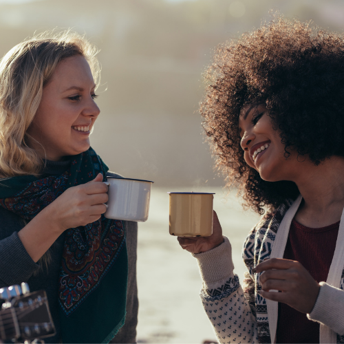 Picture of two women holding coffee mugs, smiling and enjoying the time at the beach.