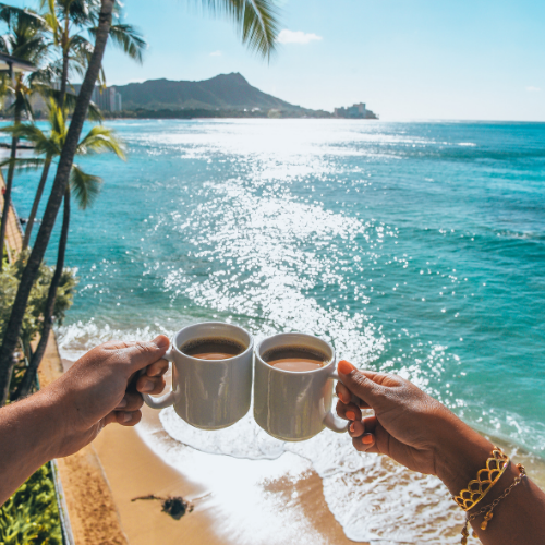 A photo of a couple toasting each other with a cup of coffee from a height overlooking a beach with lapping ocean waves
